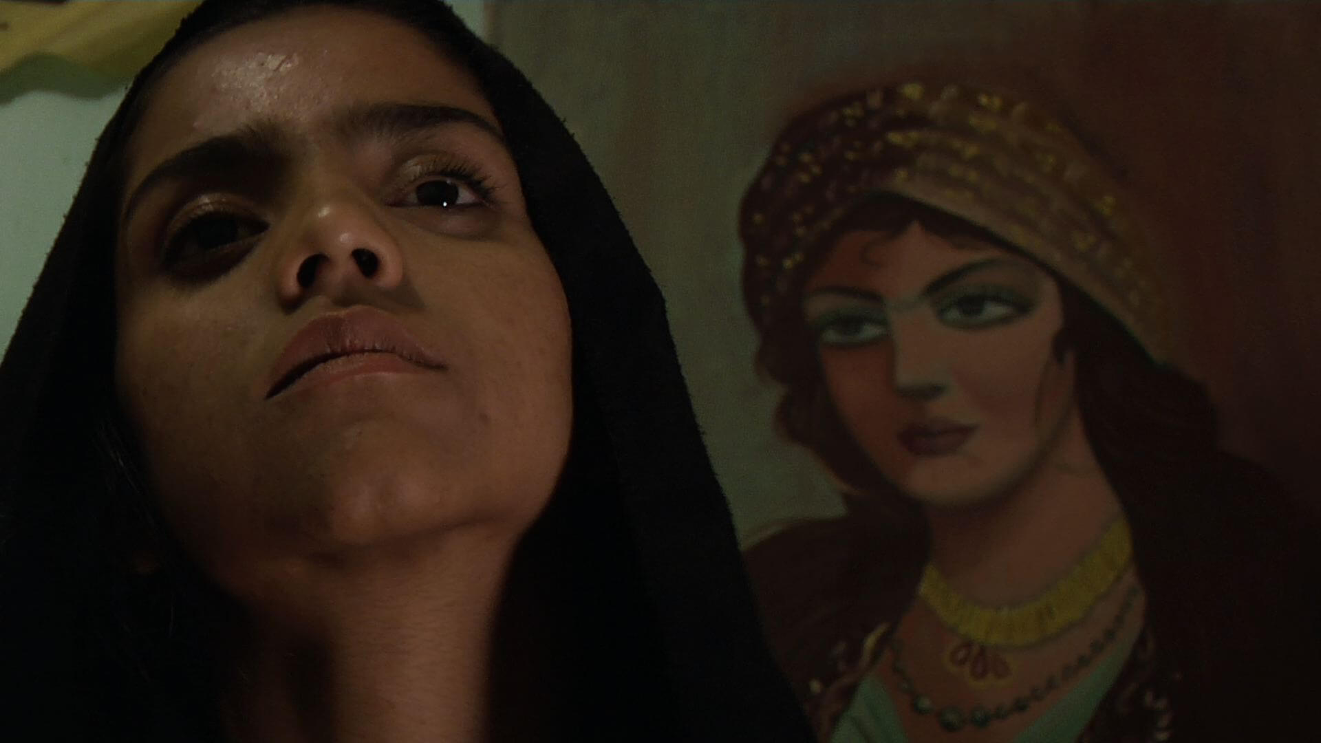 Still from Sonita. Sonita in front of the camera tilts her chin up, behind her there is a mural of a woman.