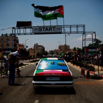 Still from Speed Sisters. Five women with racing cars suites, clasp their hands to the center and on top of a helmet, all of them look at the camera. Three Palestinian flag float in the air, in front of the finish line sign.
