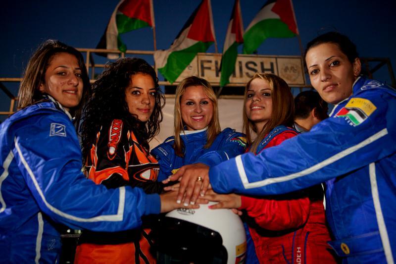 Still from Speed Sisters. Five women with racing cars suites, clasp their hands to the center and on top of a helmet, all of them look at the camera. Three Palestinian flag float in the air, in front of the finish line sign.