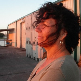 Still from Texas Gold. A photo of the side of Diane Wilson. She stands in a parking lot and looks off into the distance.