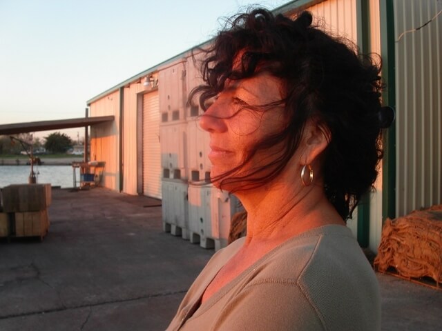 Still from Texas Gold. A photo of the side of Diane Wilson. She stands in a parking lot and looks off into the distance.