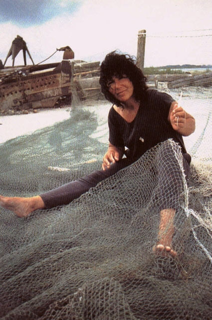 A photo of Diane Wilson sitting on the ground with a fishing net draped over her lap.