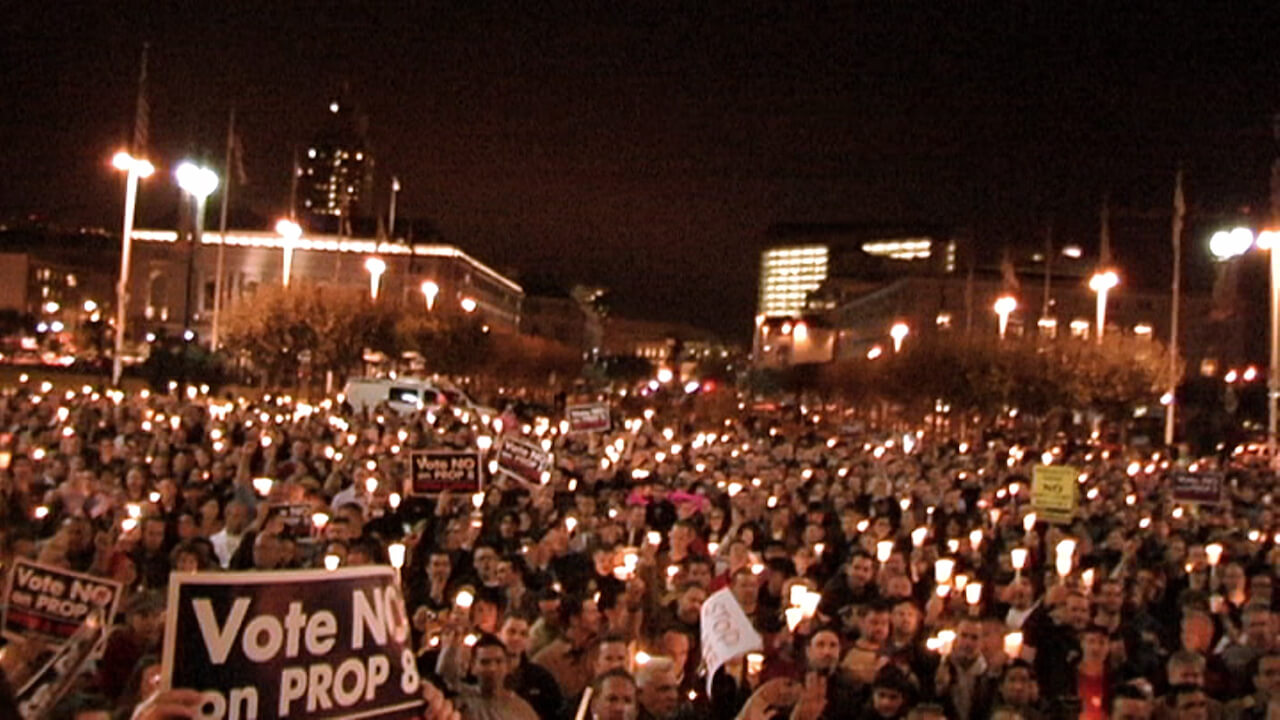 A still from The Campaign. A wide shot of a large group of people gathered during the night time. They are holding a vigil, many people hold small lights. Some signs are visible, one reads "Vote NO PROP 8".
