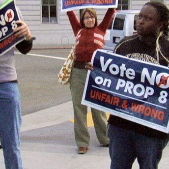 A still from The Campaign. Three people stand in the middle of the street holding signs that read "Vote NO on PROP 8: UNFAIR & WRONG".