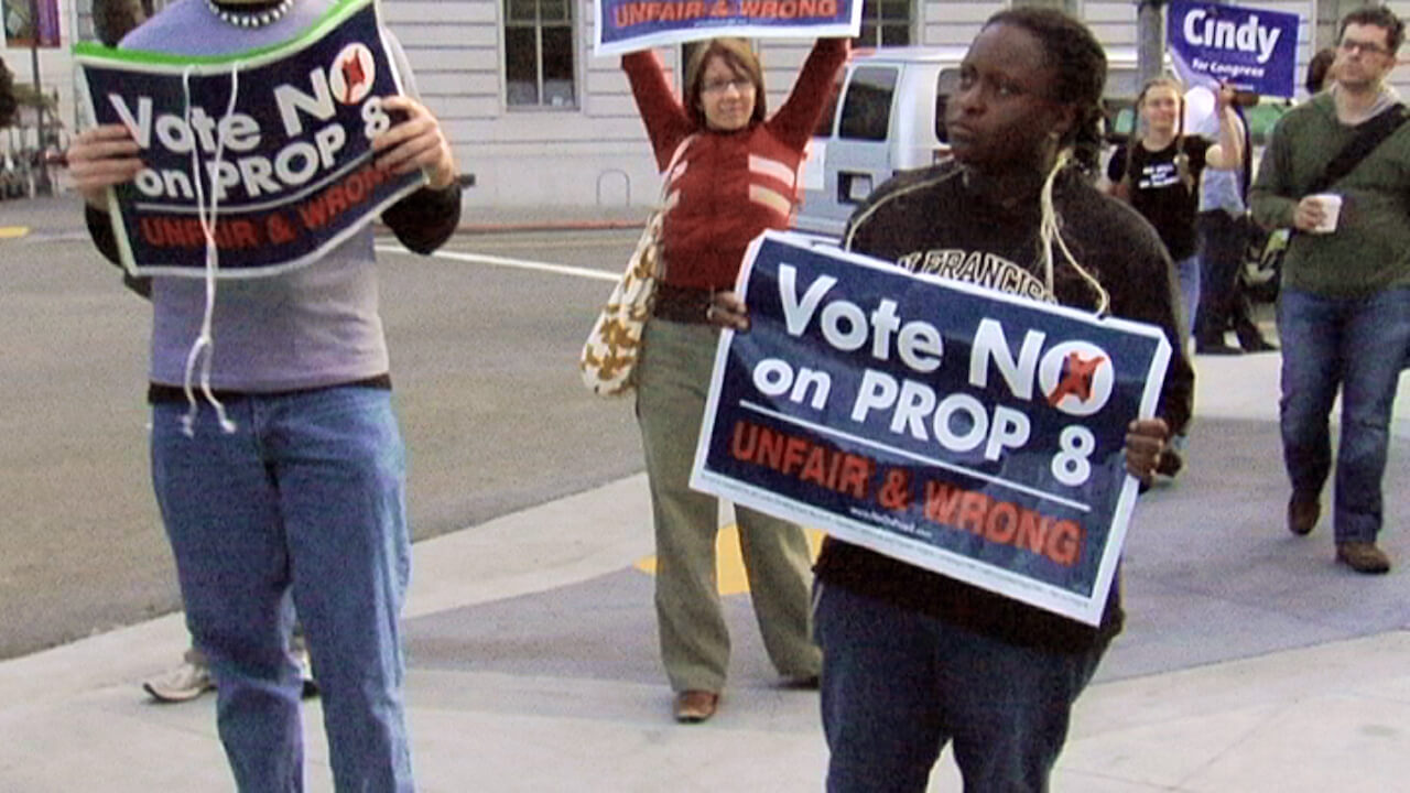 A still from The Campaign. Three people stand in the middle of the street holding signs that read "Vote NO on PROP 8: UNFAIR & WRONG".