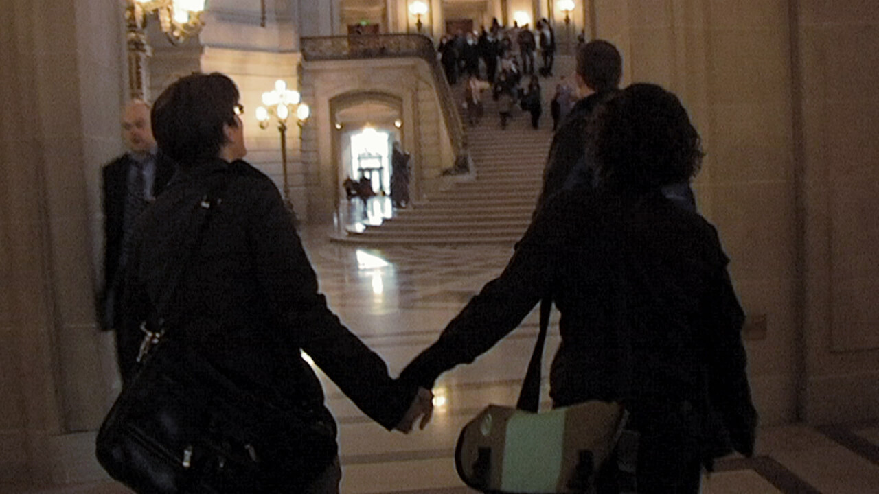 A still from The Campaign. Two people hold hands and walk through a large hall. A grand staircase sits behind them, in the distance with a group of people gathered at the top.