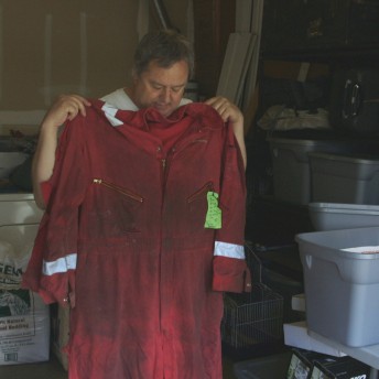 A still from The Great Invisible. A person holds a pair of dirtied red coveralls against the front of their chest and looks down at the clothing.