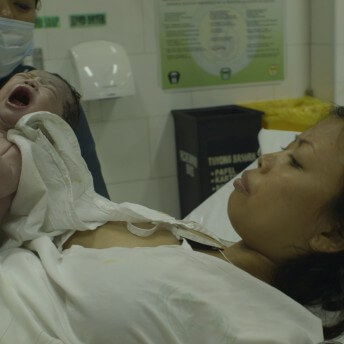 Still from Motherland. A medical professional is handing a crying newborn baby to a woman lying in a bed.