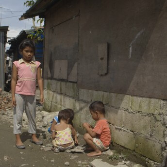 Still from Motherland. Three small children are sitting on the ground next to a building while a fourth, and older, child stands beside them looking at the camera. Another young girl walks in the background past building debris and rubble.