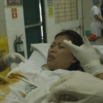 Still from Motherland. A woman surrounded by medical professionals is lying in a hospital bed, in distress.
