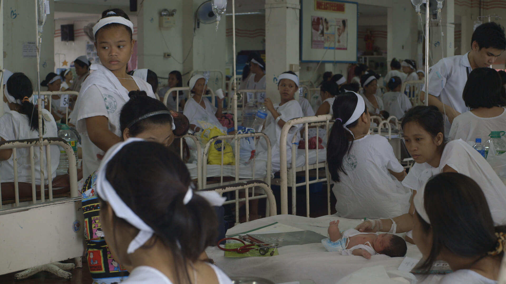 Still from Motherland. A hospital ward with many beds filled with women in various positions–standing, sitting, and talking with one another. In the center, an infant is lying on an empty bed with women surrounding them.