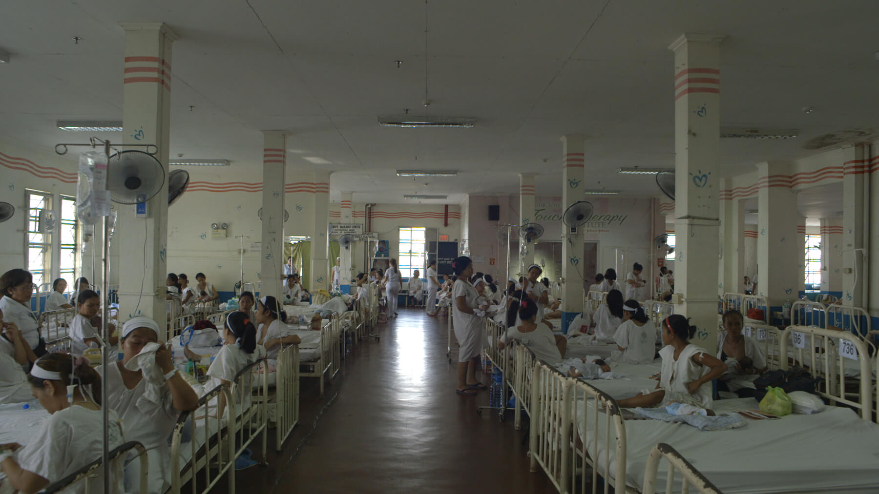 Still from Motherland. A hospital ward with many beds filled with women in various postions - standing, sitting, and talking with one another.