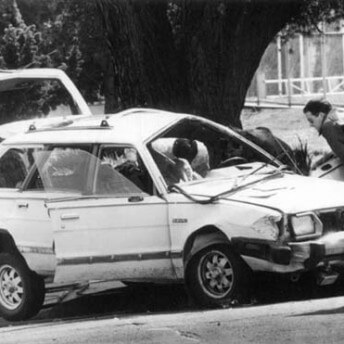 A still in black and white from The Forest for the Trees. Two people stand next to a crashed car, looking inside it.