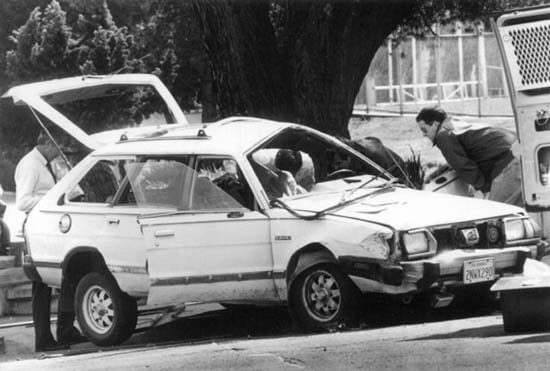 A still in black and white from The Forest for the Trees. Two people stand next to a crashed car, looking inside it.