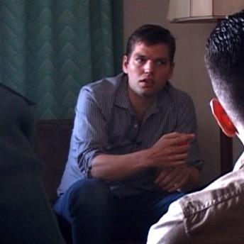 Still from The List. Two people sit in a room facing each other. The person in the front faces their back towards the camera and is facing the other person in the room directly. The other person faces the camera, leaning forward.