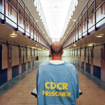 Still from The Return. A person stands in the middle of a large hallway. There are two floors of walls lined with doors. They wear a light blue jumpsuit that reads "CDCR PRISONER" on the back. They face away from the camera.
