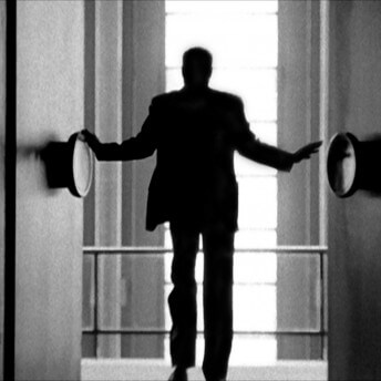 Still from Watchers of the Sky. A man in a suit stands in between two doors with large, circular knobs facing away from the camera. The man is blurry, and out of focus.