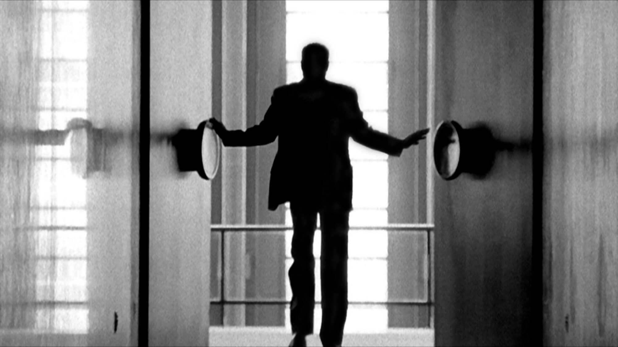 Still from Watchers of the Sky. A man in a suit stands in between two doors with large, circular knobs facing away from the camera. The man is blurry, and out of focus.