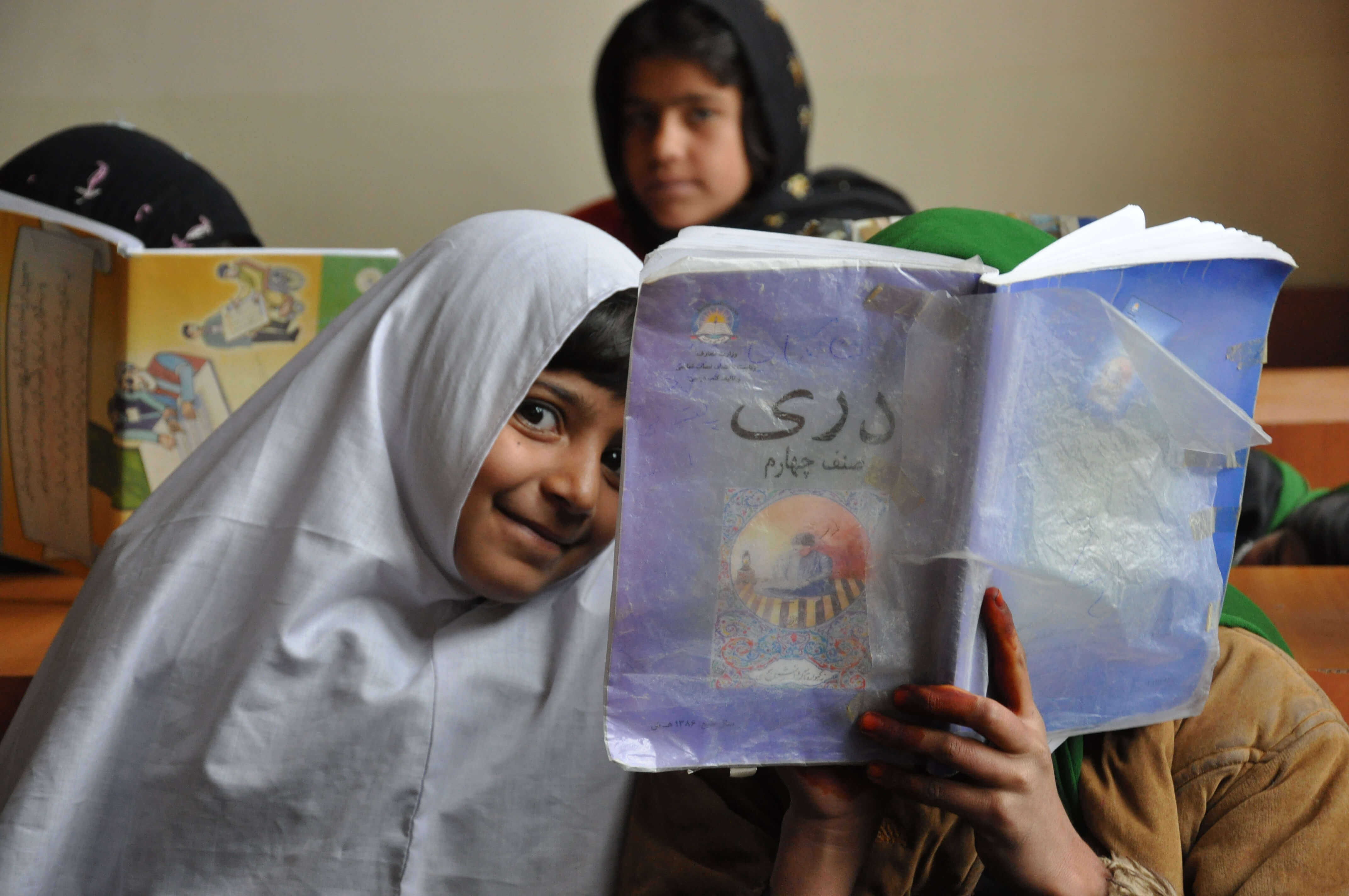 A young girl wearing a head covering looks playfully at the camera. Beside her is someone holding up a book to their face. Another girl who is out of focus looks at the camera in the background. Color photograph.