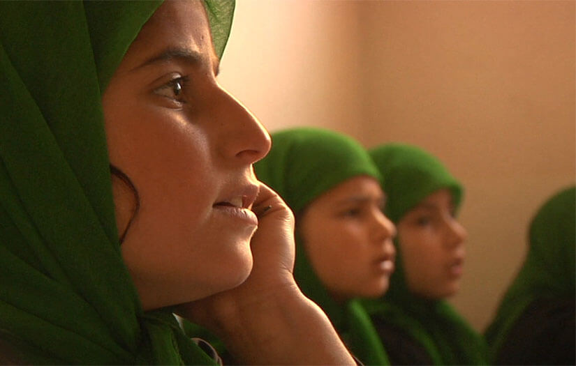 A girl wearing a head covering looks off into the distance with her hand touching her face. Two girls wearing matching head coverings are in the background out of focus. Color photograph.