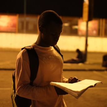 A young man is walking in the night, with a backpack on his back, and holding a notebook open in his hands.