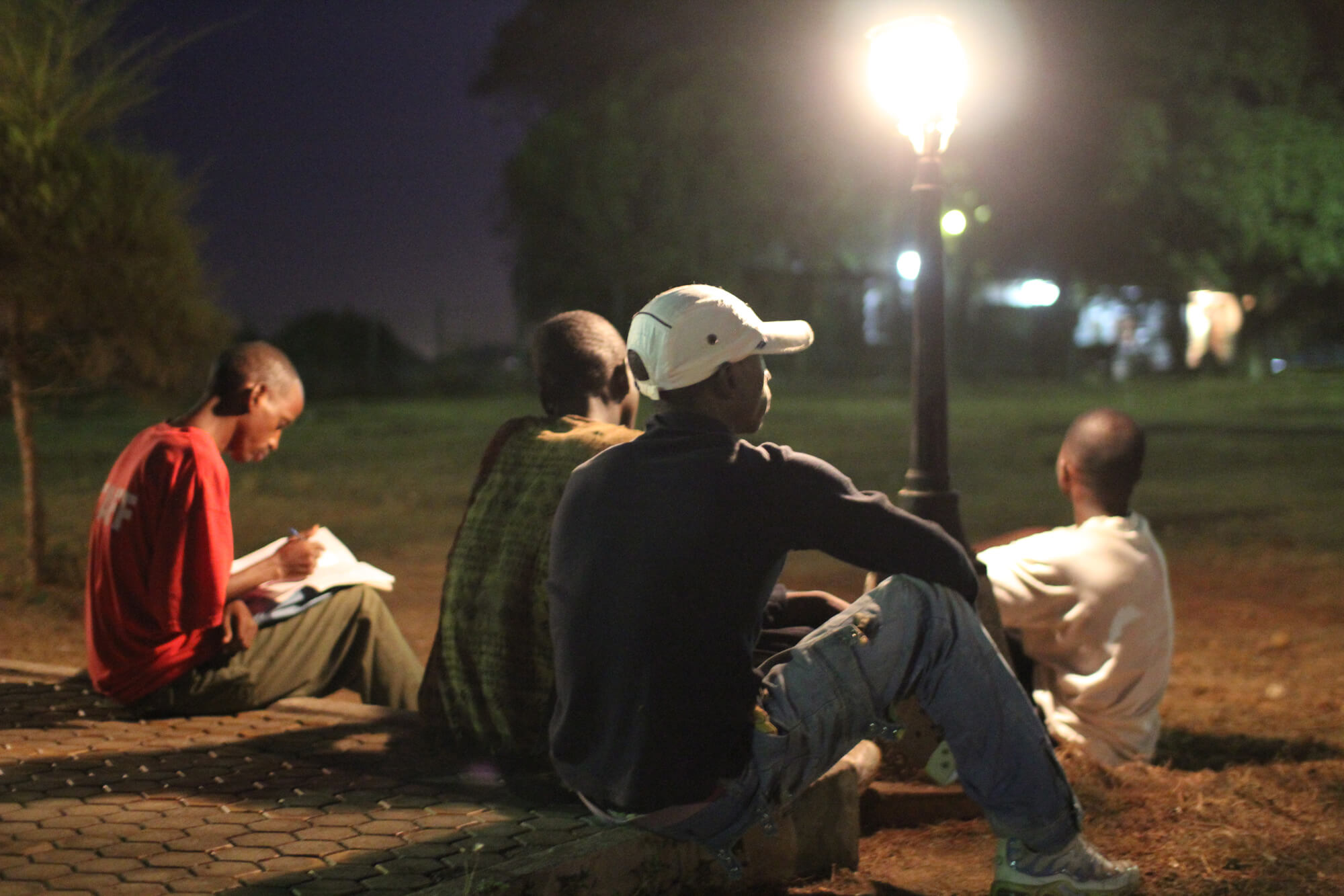 A group of young men are sitting in a park, there is a street light that is illuminating them.