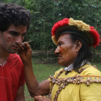 Still from Oil & Water. Two men are standing outside, facing eachother. One man, dressed in Cofan tribe traditional clothing is painting red symbols on the other's face.
