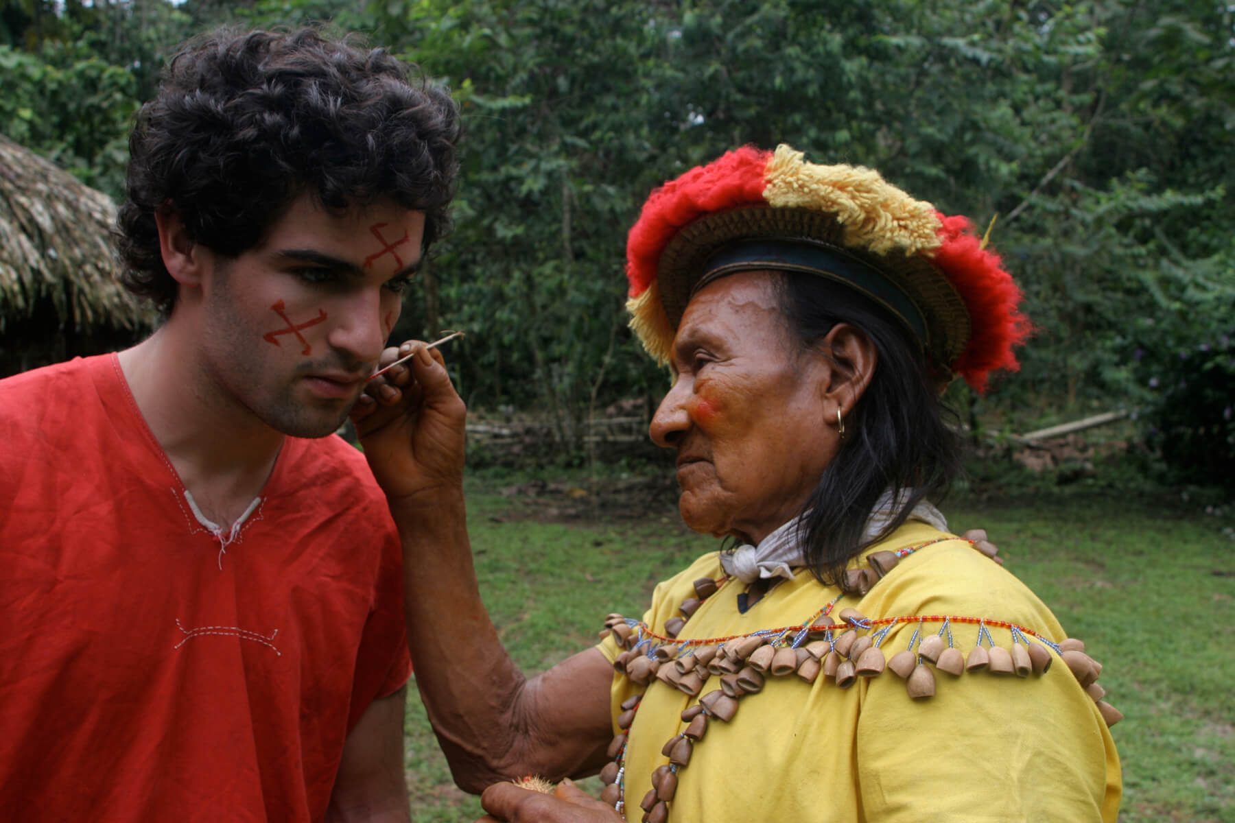 Still from Oil & Water. Two men are standing outside, facing eachother. One man, dressed in Cofan tribe traditional clothing is painting red symbols on the other's face.