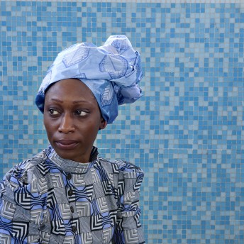 A person wearing a blue head scarf and a patterned shirt looks off into the distance. The background is a blue tiled wall. Color photograph.
