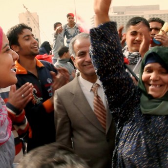 Still from Words of Witness. A group of people smile and gather outside. One woman, wearing a green hijab and black patterned top, holds up her arm.