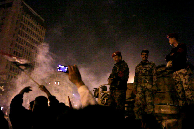 Still from Words of Witness. Two soldiers stand over a crowd of people at night. One person holds a cell phone in the air. Smoke is in the background.