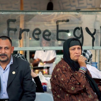 Still from Words of Witness. A man and a woman in a hijab stand in front of a glass fixture. Spray-painted in black on the glass are the words "Free Egypt."