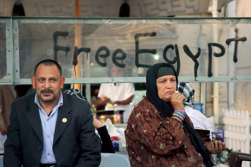 Still from Words of Witness. A man and a woman in a hijab stand in front of a glass fixture. Spray-painted in black on the glass are the words "Free Egypt."