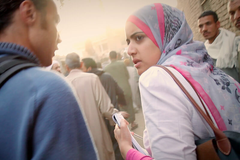 Still from Words of Witness. A woman in a white top and a gray, pink, and black hijab looks over her shoulder. A man stands beside her wearing a blue shirt.