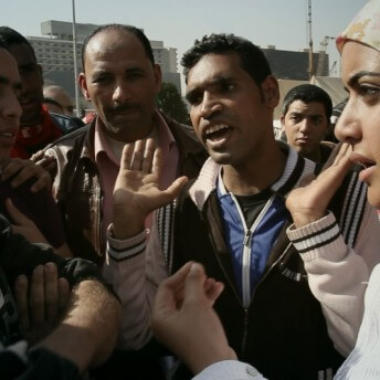 Still from Words of Witness. A group of men and a woman in a hijab stand closely in a circle outside. One man holds his hands up by his shoulders and wears a black and white jacket.