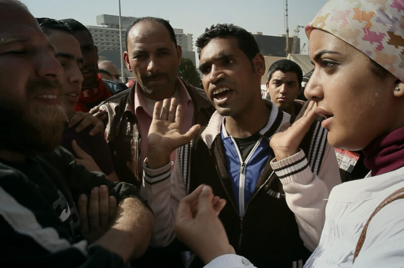 Still from Words of Witness. A group of men and a woman in a hijab stand closely in a circle outside. One man holds his hands up by his shoulders and wears a black and white jacket.