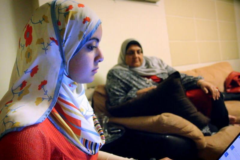 Still from Words of Witness. Two women sit on a sofa. One wears a white top and a white hijab with red flowers. The other wears a gray hijab and a top.