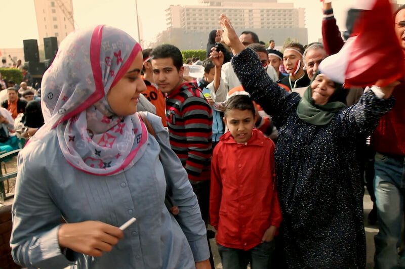 Still from Words of Witness. A crowd of people stands in the streets with their arms up. One woman, wearing a gray top with a gray, pink, and black hijab, looks in the direction of a woman with her arm up. A young boy in a red top is standing beside her.