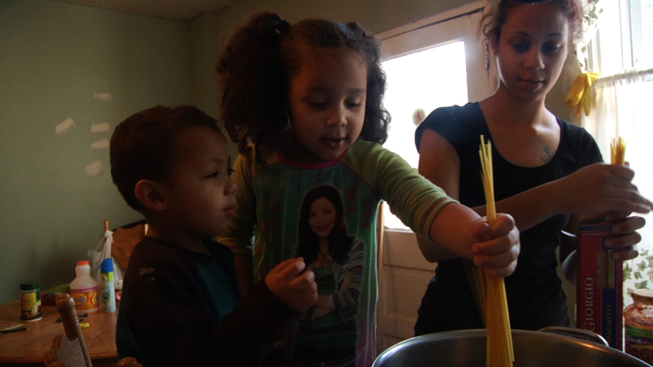 Still of A Place at the Table. A young woman is cooking spaghetti with a child and a toddler; they are putting the pasta in a pan.