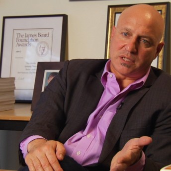 Still of A Place at the Table. Medium shot of a bald man wearing a purple buttoned shirt and a blazer; his hands are in a position as if he was explaining something. In the back, there are frames with awards and pictures.