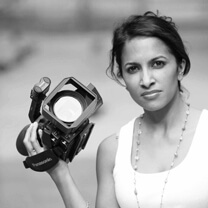 Shalini Kantayya holding a camera with an extended arm and looking directly at the camera. She wears a white tank top and a necklace, with dark hair pulled up in a bun. Portrait in black & white.