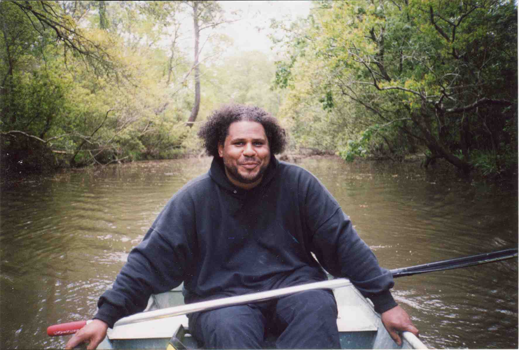 A man wearing a black hoodie and pants is rowing in a boat.