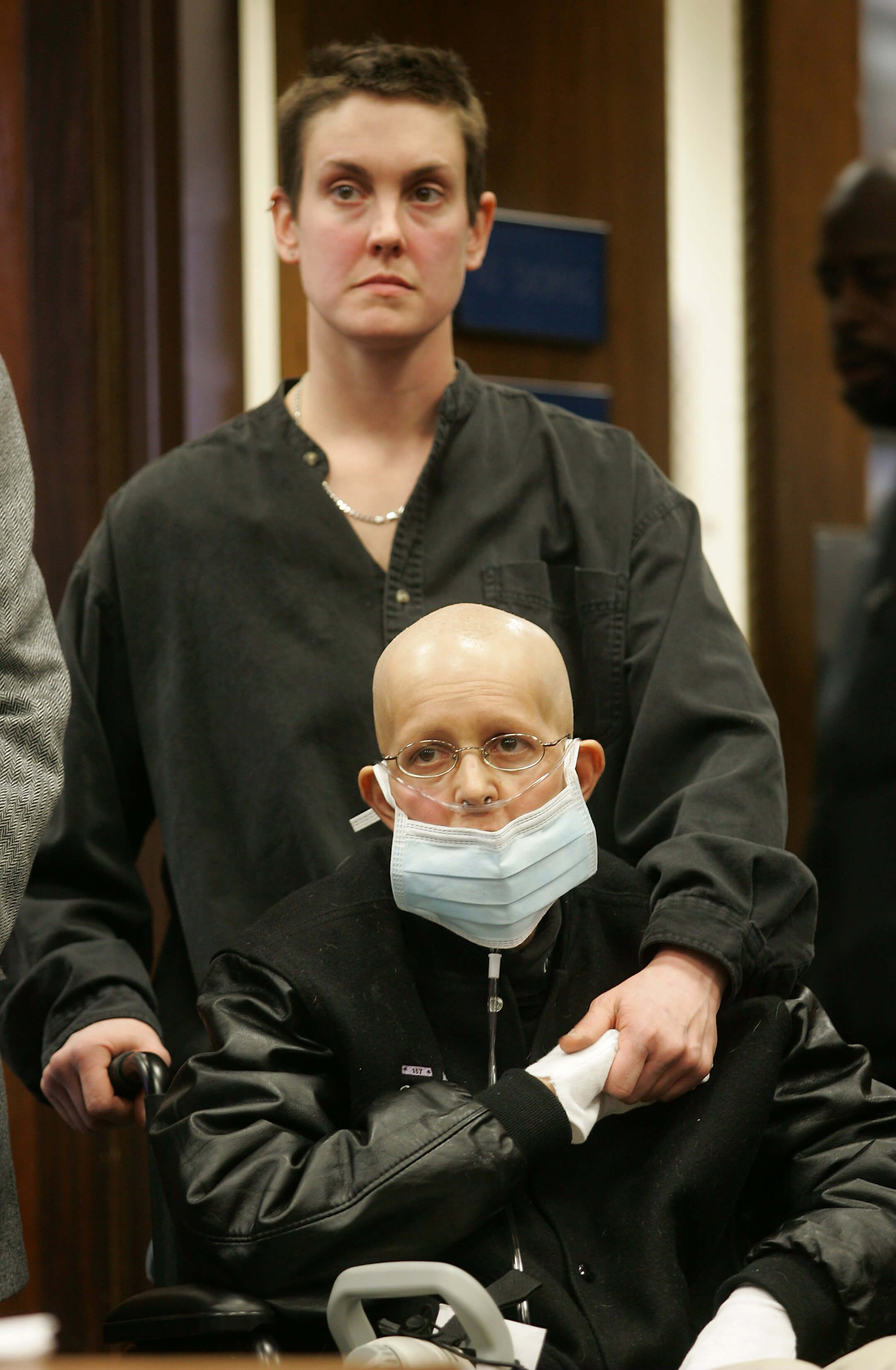 Still from Freeheld. A woman in a black letterman jacket is sitting in a wheechair. She is bald, wears silver-rimmed glasses, a nasal breathing tube, a mask over her mouth, and white gloves. She holds the hand of a short-haired woman who is standing behind her and is holding onto her wheelchair. Both women are looking at a point off-screen.