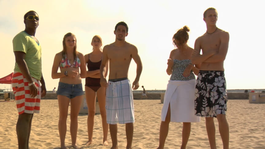 Still from Home Again. A group of six adolescents in beachwear are standing next to each other on sand. The majority are looking in the same direction offscreen.