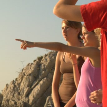 Still from Home Again. A group of three adolescent girls seen in profile. One girl is pointing in front of her, and the girl to her right is looking in the direction that she is pointing. Behind them, there is a rocky cliff, and further in the distance is seen water and a cityscape.
