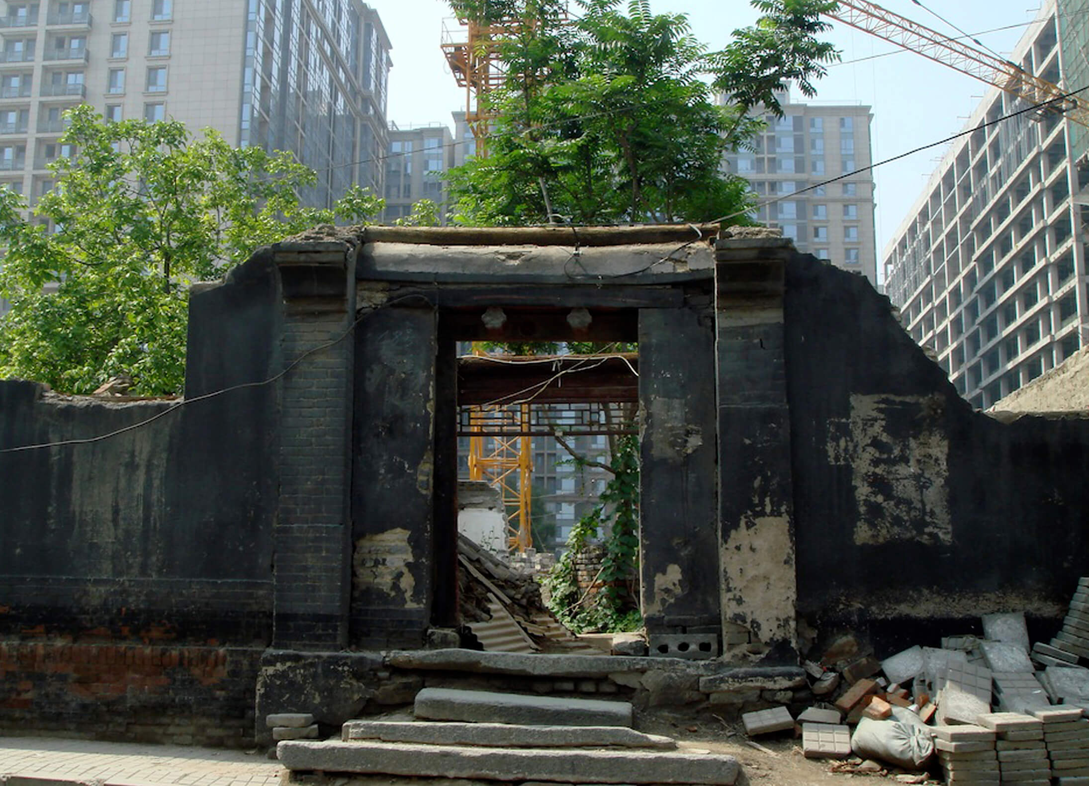 Still from Missing Home: The Last Days of the Beijing Hutongs Weimin Zhang. A damaged building structure and door frame stand among other buildings, trees, and a crane.