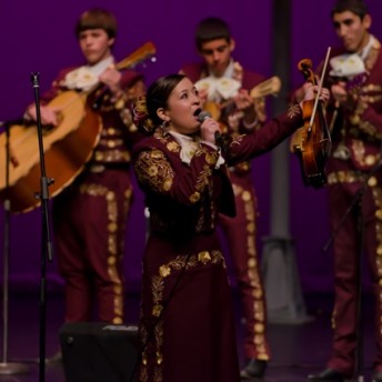 Still from Mariachi High. A woman in a purple Mariachi suit is holding up a violin and bow in her left hand and a microphone to her mouth with her right hand. Behind her are some band members playing instruments.