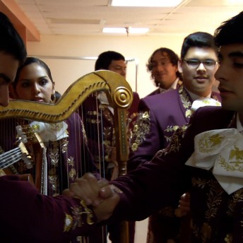 Still from Mariachi High. Two young men in the foreground shake hands in front of a young woman holding a harp among other band members in mariachi suits. They are gathered backstage.