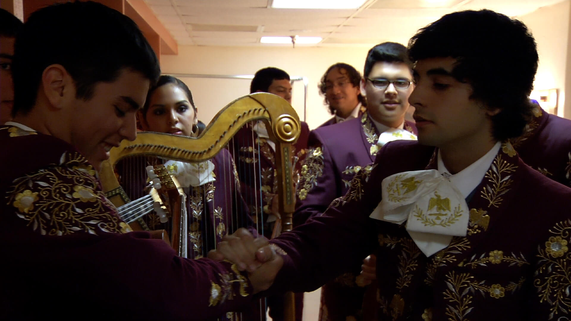 Still from Mariachi High. Two young men in the foreground shake hands in front of a young woman holding a harp among other band members in mariachi suits. They are gathered backstage.