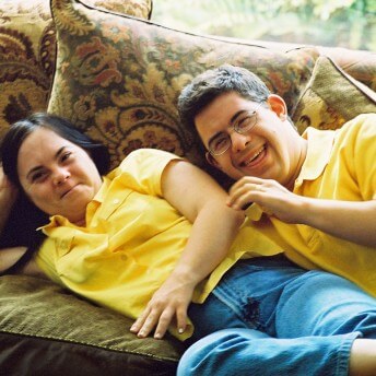 Still from Monica & David. Two people wearing yellow polo shirts and jeans are laying next to eachother on a couch with floral pillows. They are both smiling.